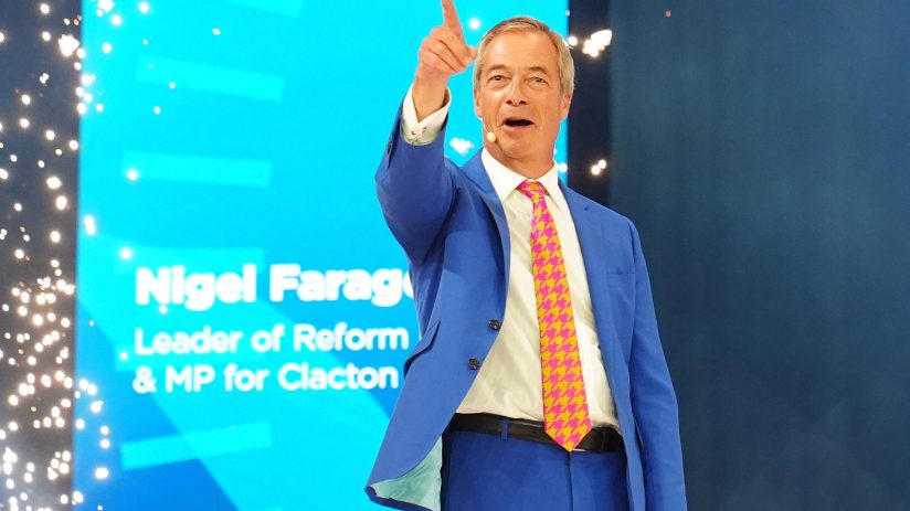 Nigel Farage speaking at the Reform UK conference, pointing upwards with a confident expression.