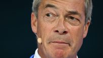 Nigel Farage close-up, looking left, with a microphone near his ear, against a dark background