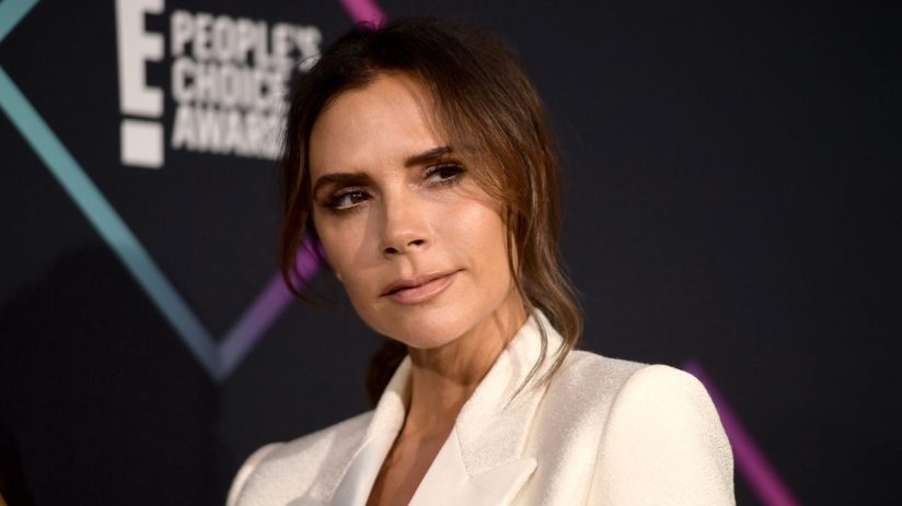 Victoria Beckham in a white blazer, looking right with a soft expression, E! Peoples Choice Awards backdrop.