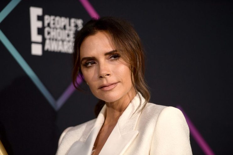 Victoria Beckham in a white blazer, looking right with a soft expression, E! Peoples Choice Awards backdrop.