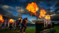 Cricket players DArcy Short and Matthew Wade walk onto the field amidst fire and fireworks at dusk.