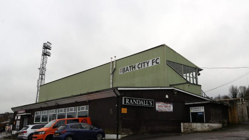 Exterior view of Bath City FC stadium with Randalls signage, parked cars, and a tall floodlight against a cloudy sky.