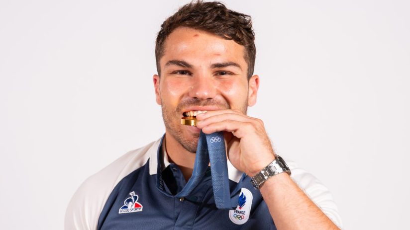 Smiling man biting a gold medal, wearing a blue and white French Olympic uniform and a watch