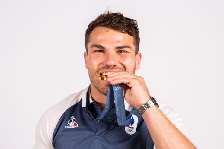 Smiling man biting a gold medal, wearing a blue and white French Olympic uniform and a watch