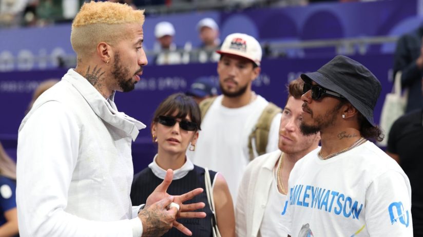 Lewis Hamilton and fencing champion Miles Chamley-Watson converse with others in a sports arena.