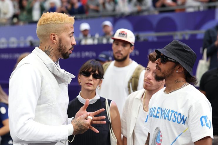 Lewis Hamilton and fencing champion Miles Chamley-Watson converse with others in a sports arena.