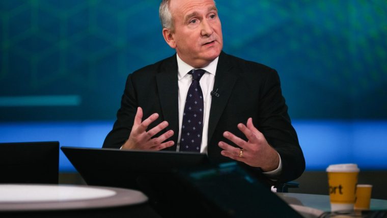 Businessman in a suit and patterned tie gesturing while speaking in a studio setting.