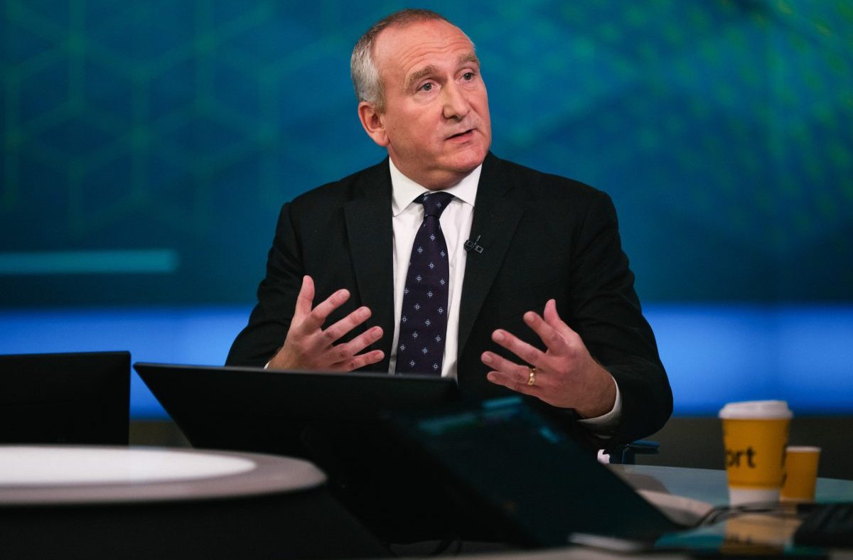 Businessman in a suit and patterned tie gesturing while speaking in a studio setting.
