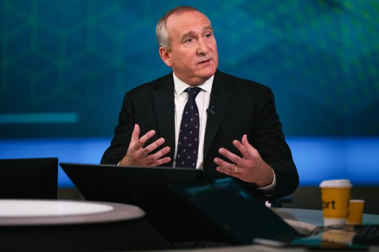 Businessman in a suit and patterned tie gesturing while speaking in a studio setting.