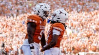 Two college football players in orange and white uniforms celebrate on a field with a crowd in the background