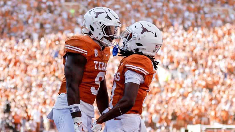 Two college football players in orange and white uniforms celebrate on a field with a crowd in the background