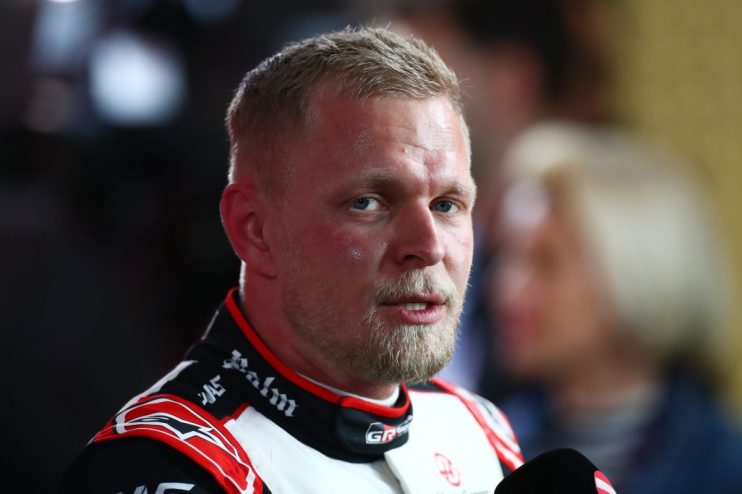 Man with blond hair and beard wearing a racing suit, speaking into a microphone at a news event.