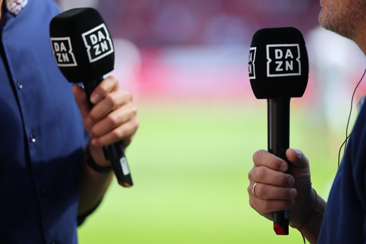 Two people hold black DAZN branded microphones, green field in background. Sports broadcasting, media coverage.