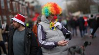Person in a rainbow clown wig and red nose using a flip phone, next to a man in a red and white bucket hat.