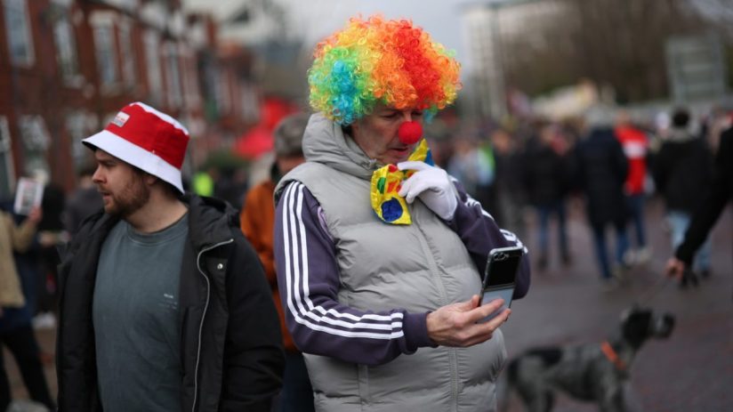 Person in a rainbow clown wig and red nose using a flip phone, next to a man in a red and white bucket hat.
