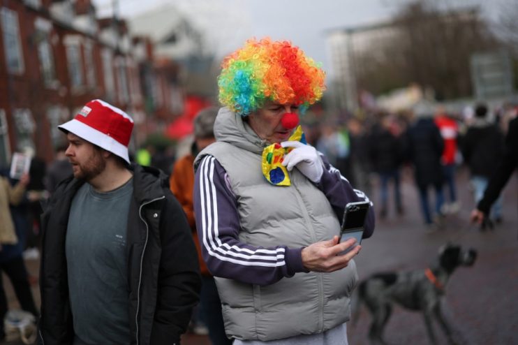 Person in a rainbow clown wig and red nose using a flip phone, next to a man in a red and white bucket hat.