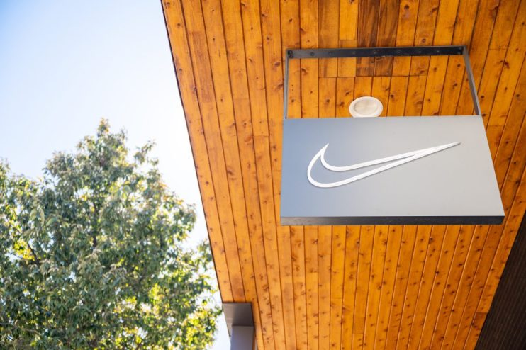 Nike swoosh logo sign hanging from a modern wooden building facade, with a green tree and blue sky in the background.