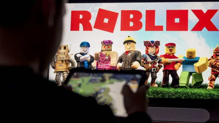 Person playing Roblox on a mobile device with the game logo and avatars displayed on a large screen