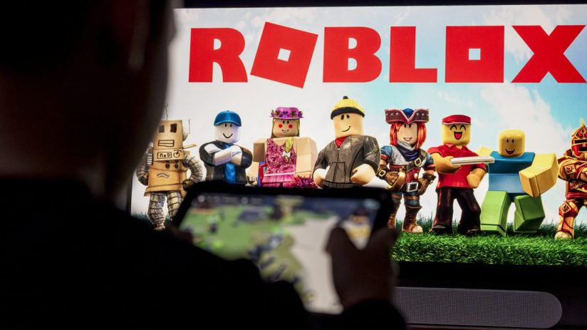 Person playing Roblox on a mobile device with the game logo and avatars displayed on a large screen