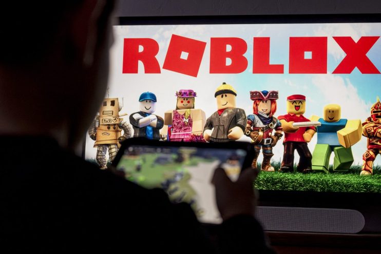 Person playing Roblox on a mobile device with the game logo and avatars displayed on a large screen