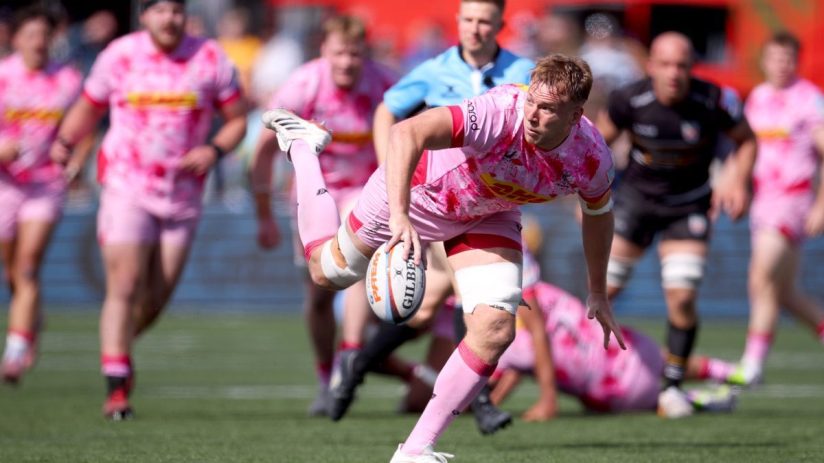 Rugby player in pink jersey with ball, surrounded by teammates on a green field during a match.