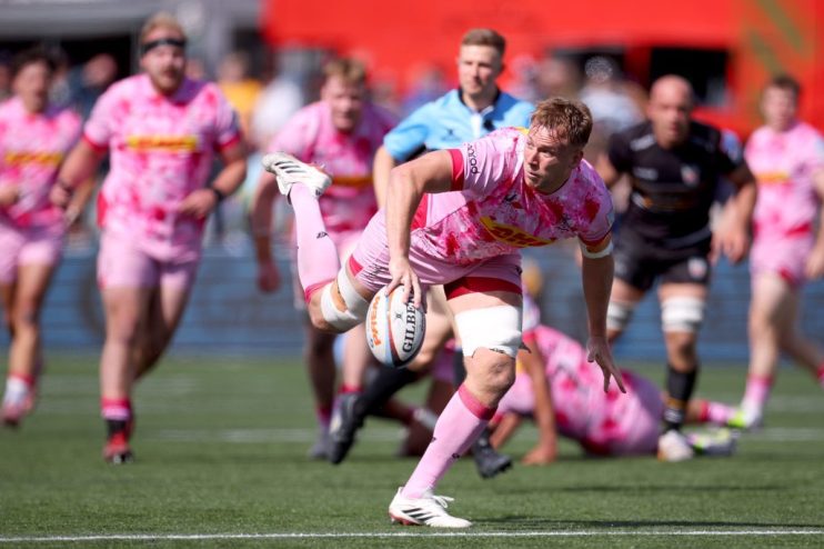 Rugby player in pink jersey with ball, surrounded by teammates on a green field during a match.