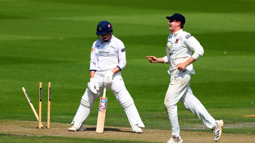 Cricket batsman looking dejected while an opposing player celebrates near fallen stumps.