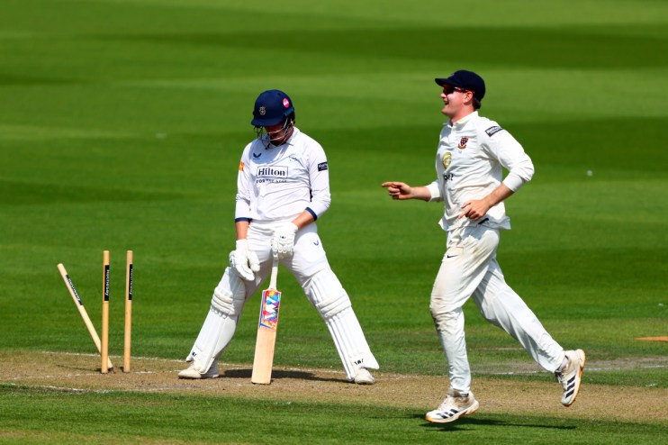 Cricket batsman looking dejected while an opposing player celebrates near fallen stumps.