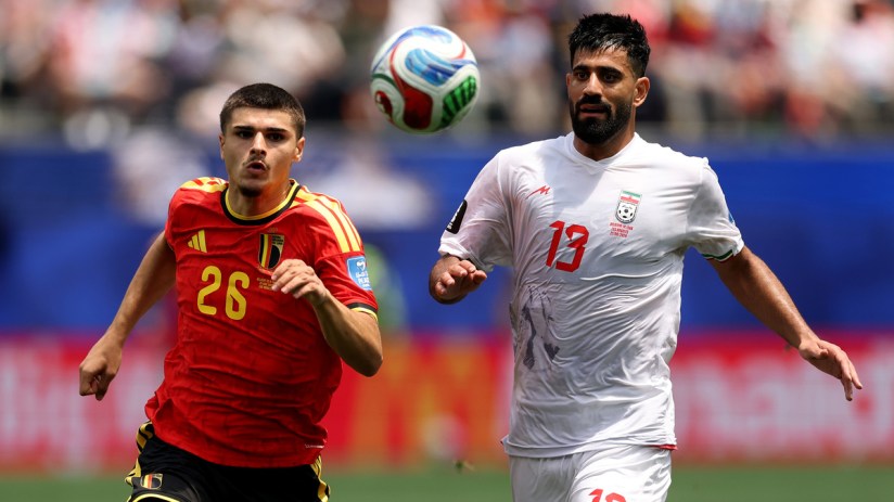 Two male soccer players, one in a red Belgium jersey and one in a white Iran jersey, compete for a ball mid-air.