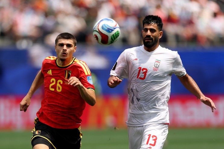 Two male soccer players, one in a red Belgium jersey and one in a white Iran jersey, compete for a ball mid-air.