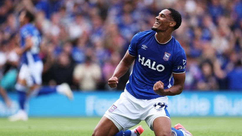 Footballer in a blue jersey celebrating on his knees on a green field with fans in the background.