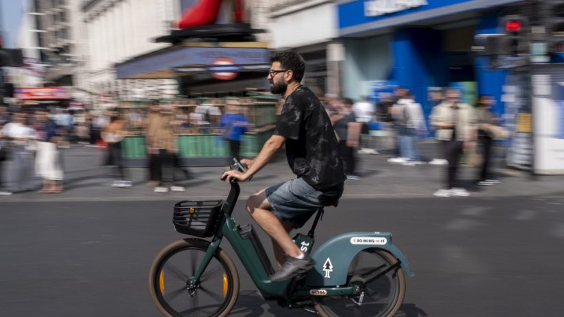 Man in glasses and beard riding a green rental e-bike on a city street with blurred background of buildings and people.