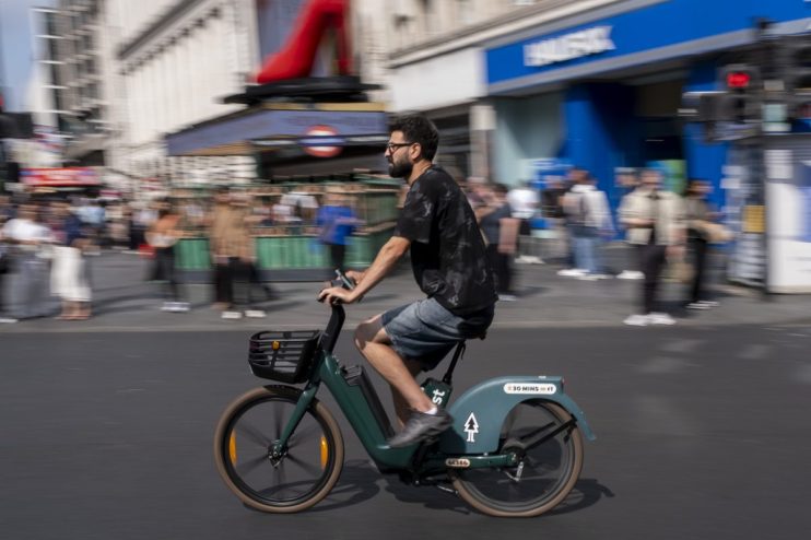Man in glasses and beard riding a green rental e-bike on a city street with blurred background of buildings and people.