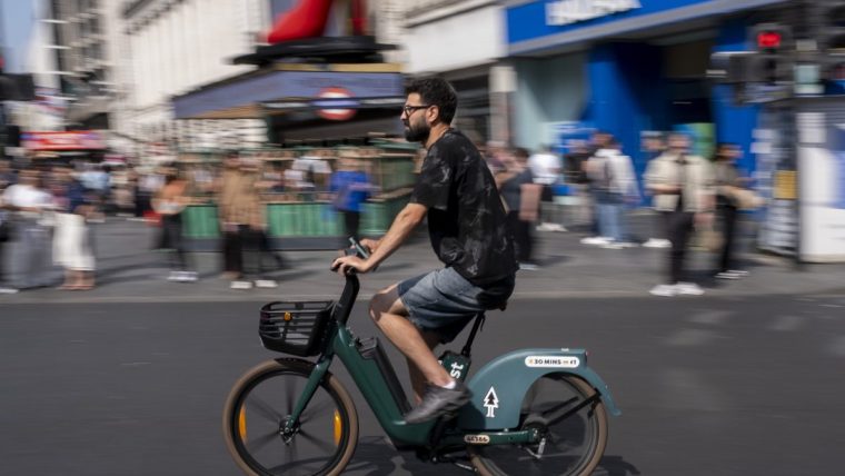Man in glasses and beard riding a green rental e-bike on a city street with blurred background of buildings and people.