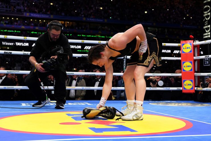 Female boxer Katie Taylor picks up her boxing gloves in the ring after a match, with a cameraman nearby.