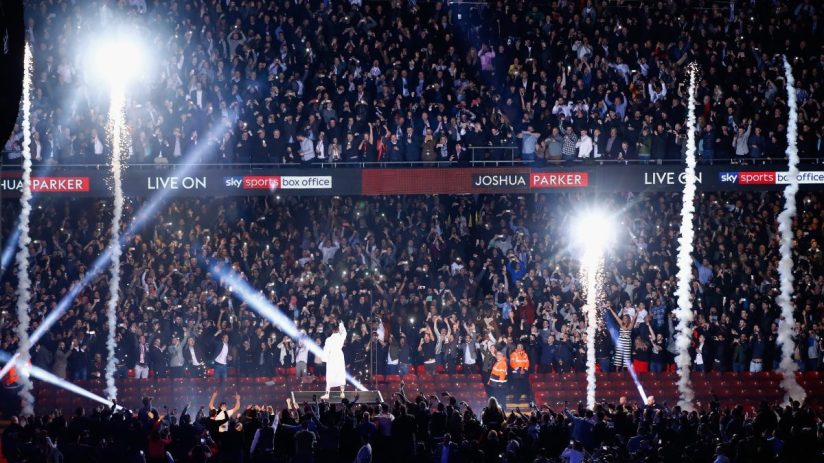 Crowd cheers with phone lights, pyrotechnics, and a performer on stage under Joshua Parker and Sky Sports Box Office signs.