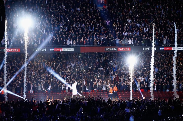 Crowd cheers with phone lights, pyrotechnics, and a performer on stage under Joshua Parker and Sky Sports Box Office signs.