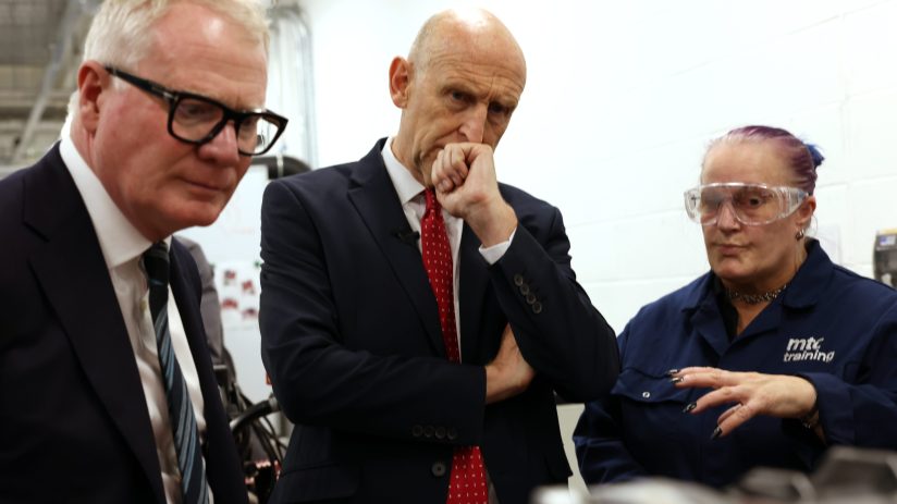 Two men in suits and a woman in safety glasses and workwear at a factory.