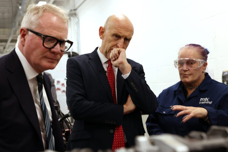 Two men in suits and a woman in safety glasses and workwear at a factory.