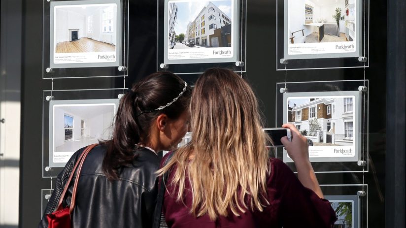 Two women view London housing property listings in a real estate agents window, one takes a phone photo.