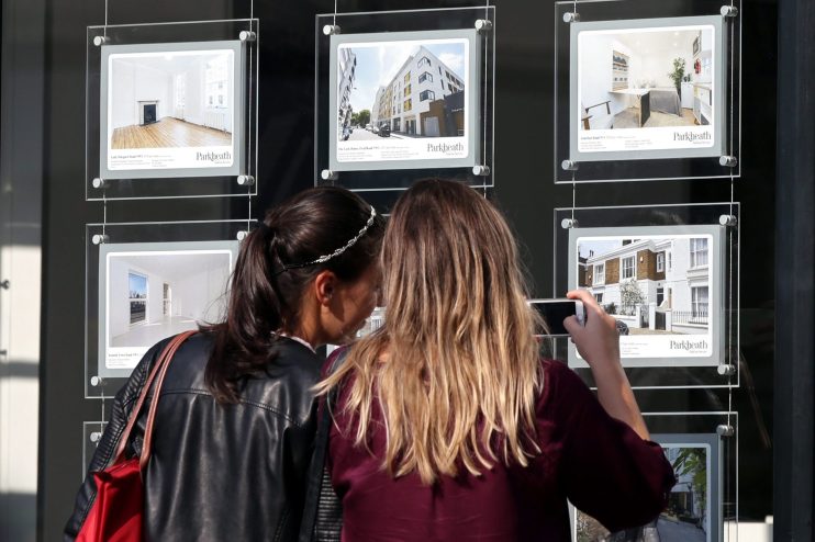 Two women view London housing property listings in a real estate agents window, one takes a phone photo.