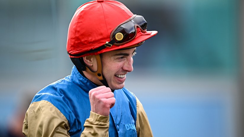 James Doyle, a jockey, in a red helmet and blue and gold silks, smiling and clenching his fist.