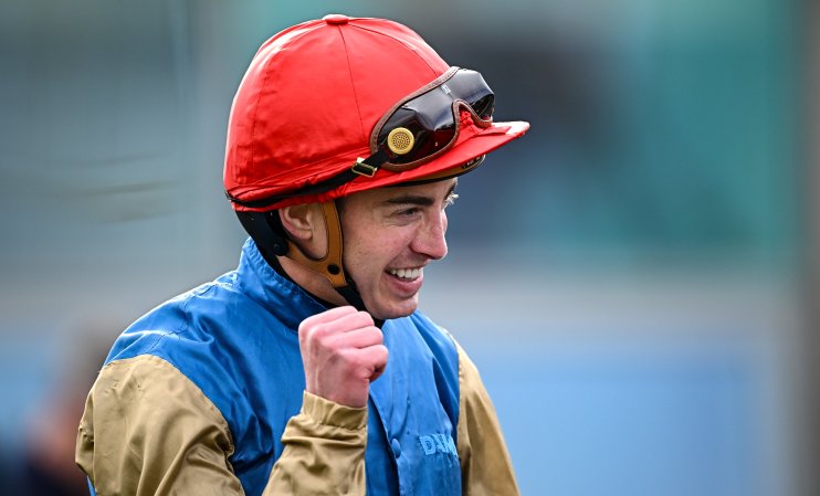 James Doyle, a jockey, in a red helmet and blue and gold silks, smiling and clenching his fist.