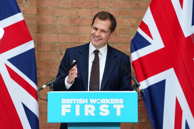Robert Jenrick speaking at a podium with British Workers First and Union Jack flags, discussing bank taxes.