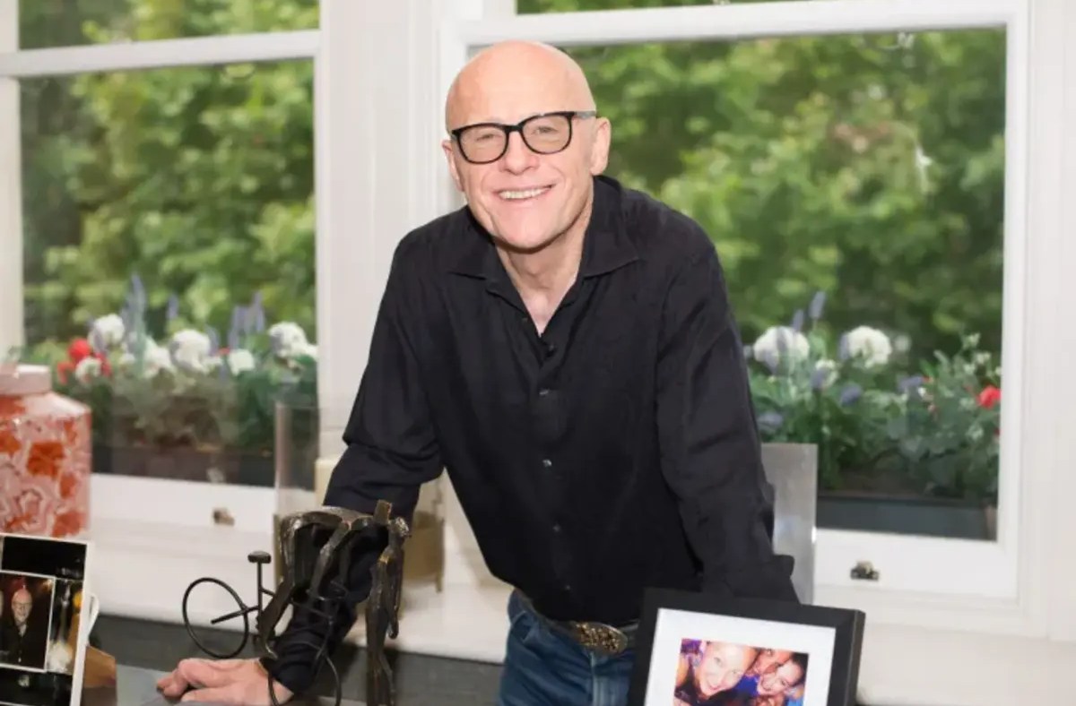 John Caudwell, bald man in black shirt and glasses, smiling, leaning on a desk with window garden background.