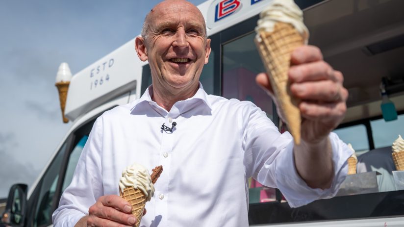 John Healey smiling, holding two ice cream cones, standing in front of an ice cream van.