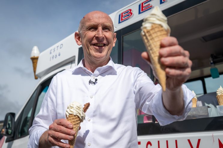 John Healey smiling, holding two ice cream cones, standing in front of an ice cream van.