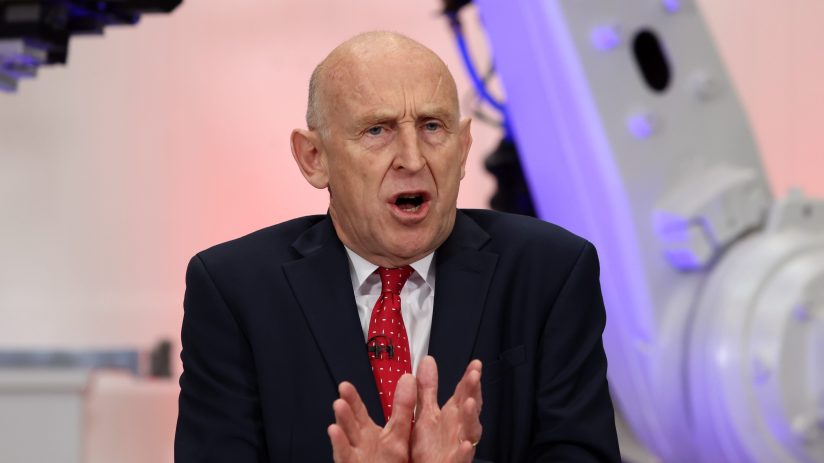 John Healey speaking at JLR, with industrial robotic arms visible in the background