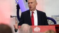 John Healey speaking at a podium with the UK Royal Coat of Arms, gesturing with his left hand during his speech.