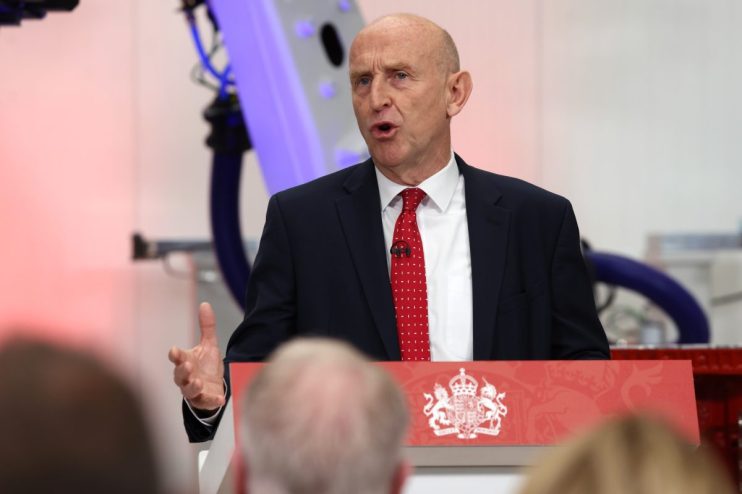 John Healey speaking at a podium with the UK Royal Coat of Arms, gesturing with his left hand during his speech.